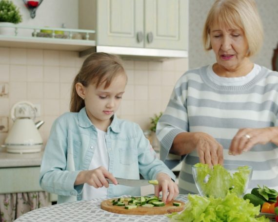 Grandma and cute child are cooking salad for lunch talking making healthy meal at home. Domestic activities, family life and happy childhood concept.